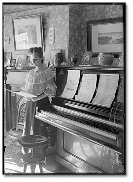 Photo: Woman looking at sheet music, posed beside a piano, [between 1898 and 1920]