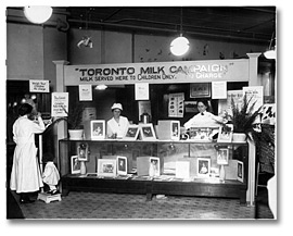 Photo: Nurses giving out free milk and weighing a child at a booth, T. Eaton Co. store, as part of the Toronto Milk Campaign, 1921
