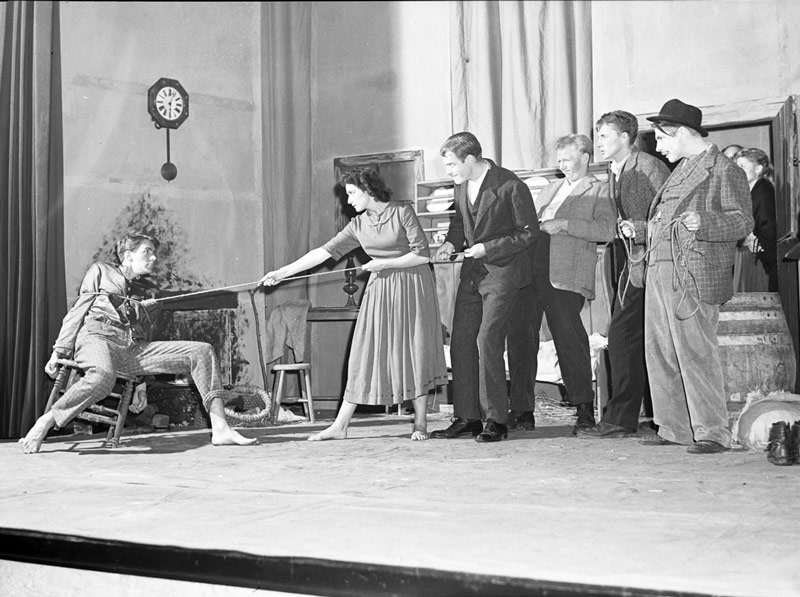 Six white actors on stage. Five actors pull on a rope around a man tied to a chair.