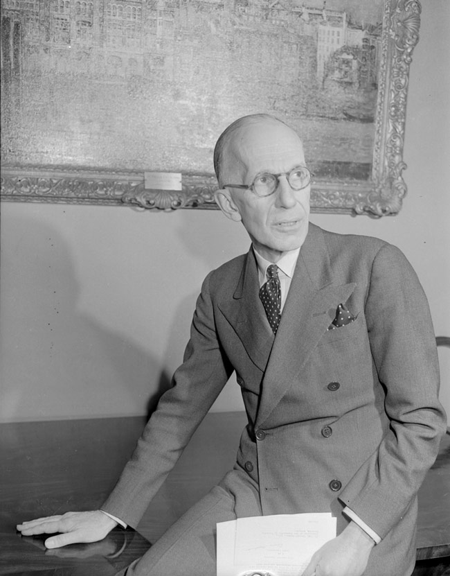 Vincent Massey, assis sur le rebord d’un meuble, portant un complet, une cravate et des lunettes et tenant un document à la main
