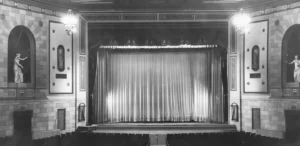 Black-and-white photo of a theatre, looking towards the stage with a drawn curtain. On the walls to either side of the stage are two niches, each containing a Classical-style sculpture of a woman gesturing towards the stage.