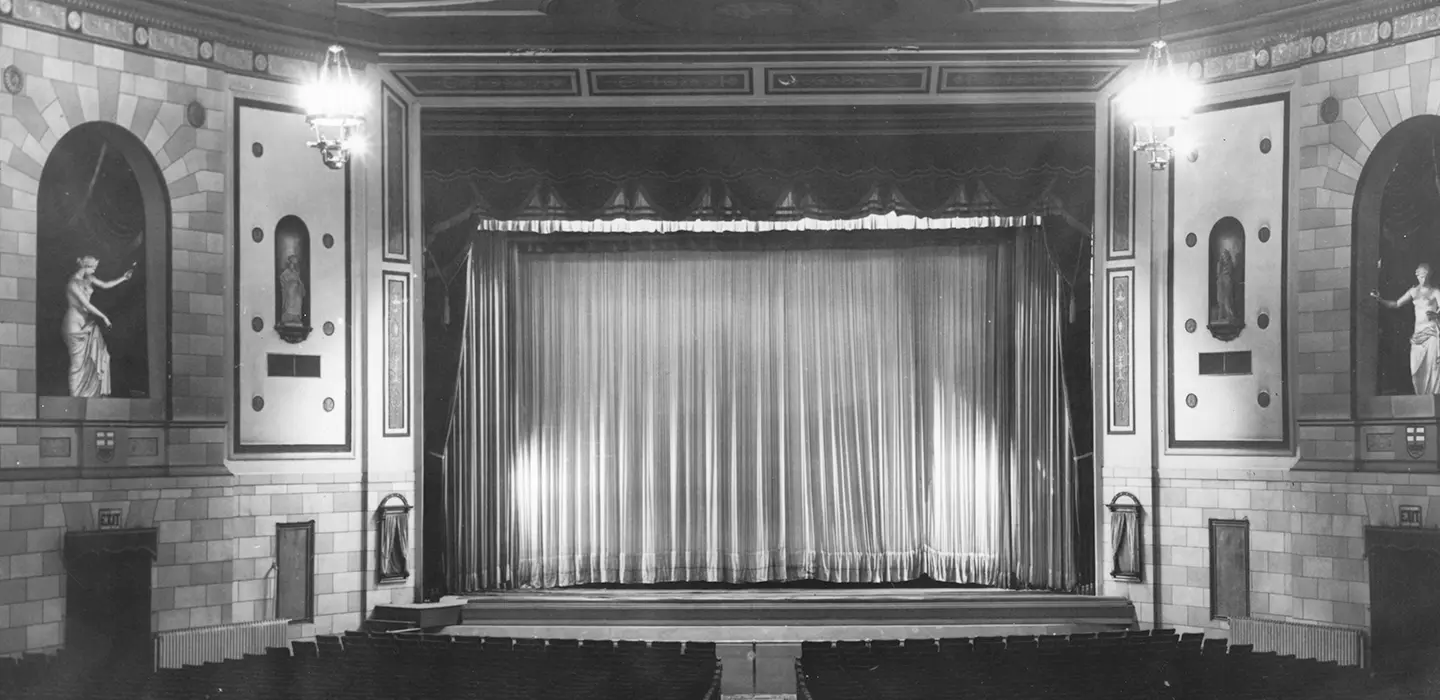 Black-and-white photo of a theatre, looking towards the stage with a drawn curtain. On the walls to either side of the stage are two niches, each containing a Classical-style sculpture of a woman gesturing towards the stage.