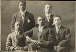 Black-and-white studio portrait photo of five young Black men in suits and ties, two of them standing and three seated in front, one holding a rolled-up piece of paper.