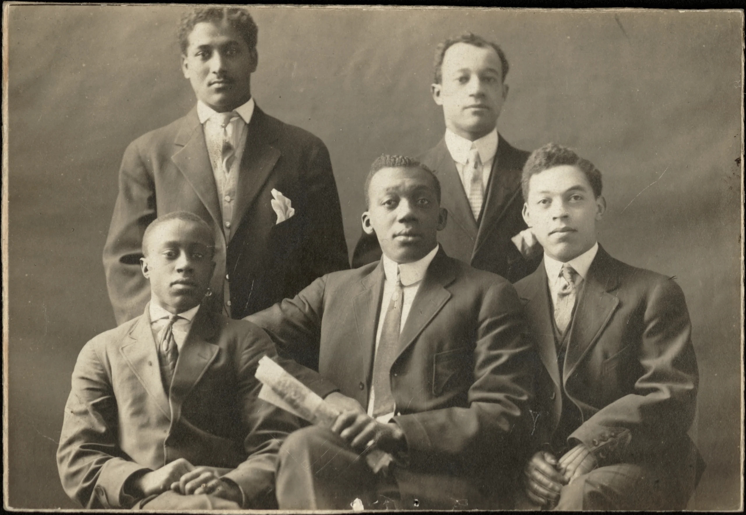 Black-and-white studio portrait photo of five young Black men in suits and ties, two of them standing and three seated in front, one holding a rolled-up piece of paper.