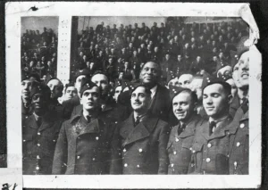 Black-and-white photo of a racially diverse group of men in military uniforms standing together with a large crowd in the background.