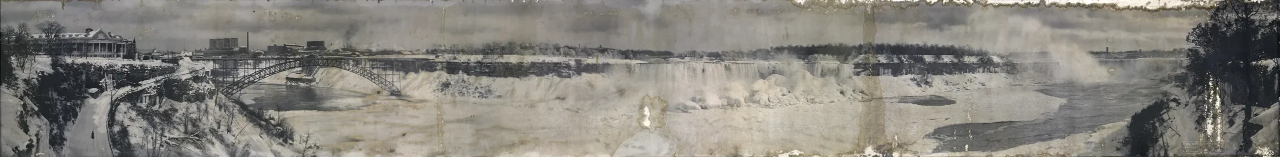 Photographie panoramique en noir et blanc de Niagara Falls en hiver, couverte de neige et de glace. À gauche, un pont enjambe la rivière; on distingue un chemin enneigé menant à un grand bâtiment au sommet d’une colline. À droite, on voit un sentier bordé d’arbres. Des déchirures et des taches d’eau sont visibles sur l’image.