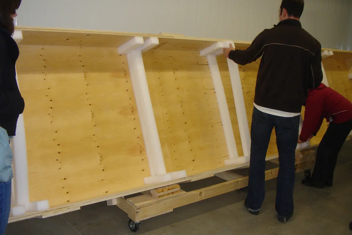 Trois personnes à côté d’une longue caisse en bois ouverte avec des bandes de mousse qui tapissent l’intérieur. La caisse est inclinée sur le côté sur un châssis en bois roulant devant un mur blanc.