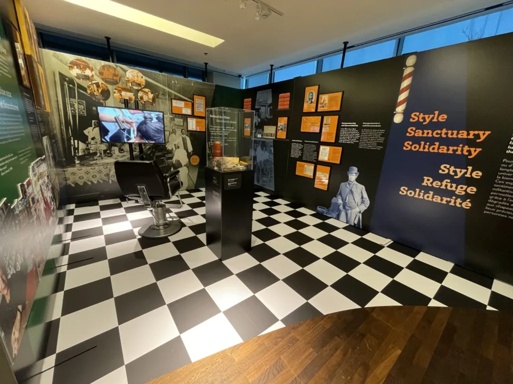 Colourful exhibit display featuring wall panels decorated with text and images. The floor is covered with a black-and-white checkered tile pattern, and a barber chair and small plinth topped with a display of barber’s tools are positioned in the centre of the space.