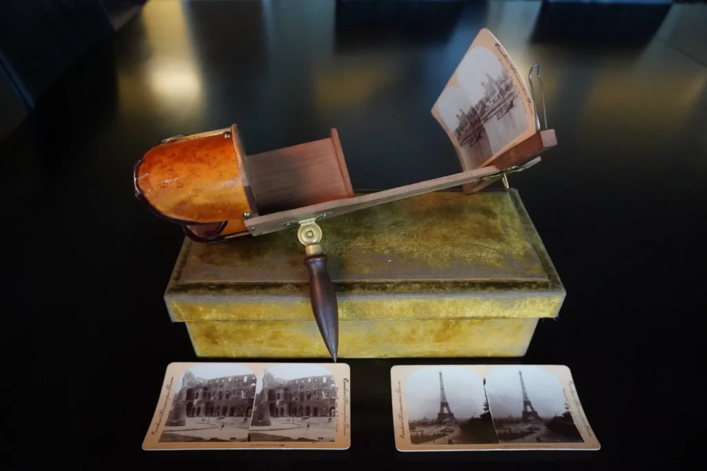 A brown, wooden viewfinder device sits on top of a worn, chartreuse velvet box. Two stereocards, with black-and-white images, are displayed in front of the box. The objects sit on a dark surface.