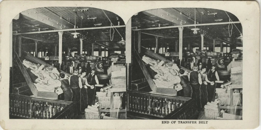 Black-and-white photographic print on card depicting the end of a large conveyor belt in a large, open building. There are carved wooden railings in the foreground and the handful of men moving goods from the belt are in waistcoats. The image appears to be duplicated, and the duplicates are side-by-side.