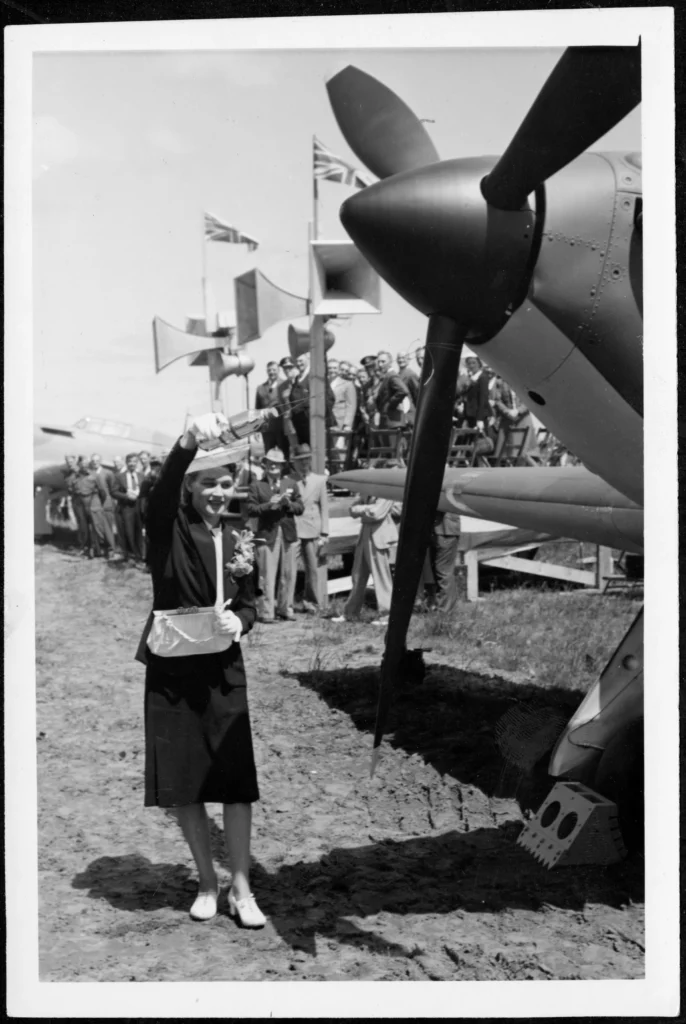 Black-and-white image of a woman standing in front of an airplane’s propeller posing as though pouring a bottle toward the plane.