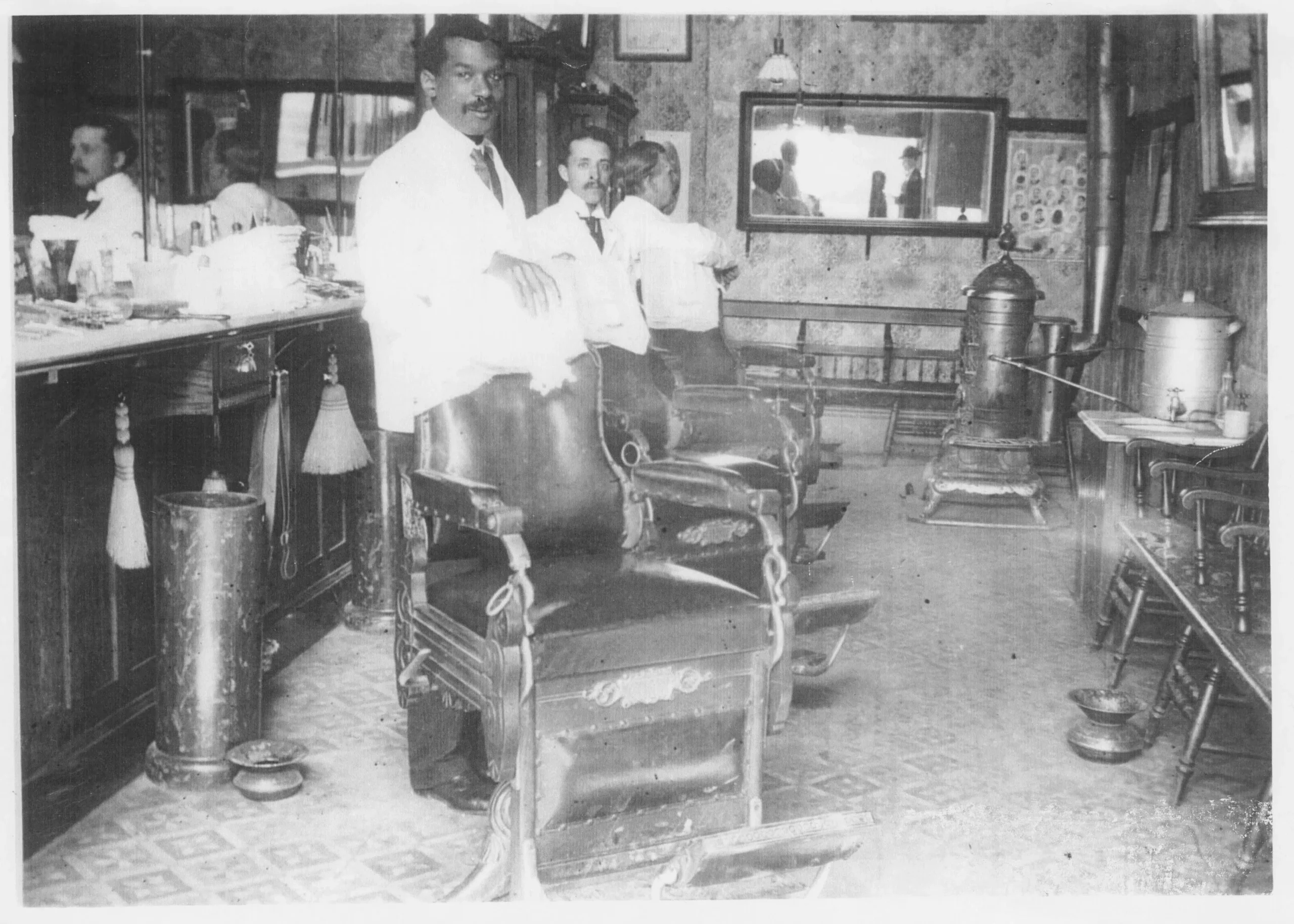 Black-and-white photo of two Black men and one white man standing in a wallpapered and floor-tiled barbershop interior outfitted with barber chairs, large mirrors, and various objects and tools related to the barbering trade. The two Black men look at the camera while the white man looks away from it, toward the shop’s back wall.