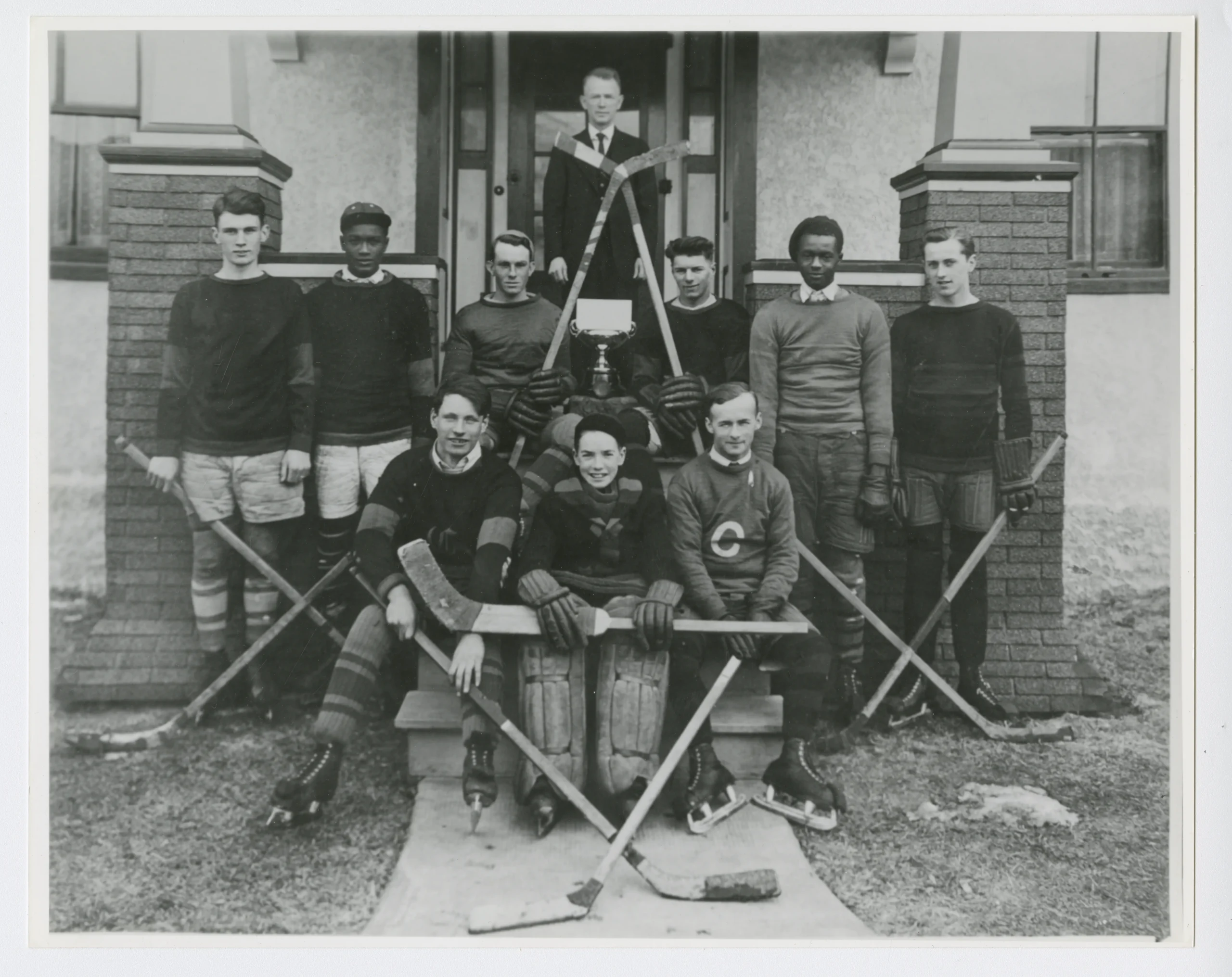 Photo portrait noir et blanc d’un groupe de jeunes hockeyeurs en uniforme, posant avec leurs bâtons devant l’entrée d’un bâtiment.