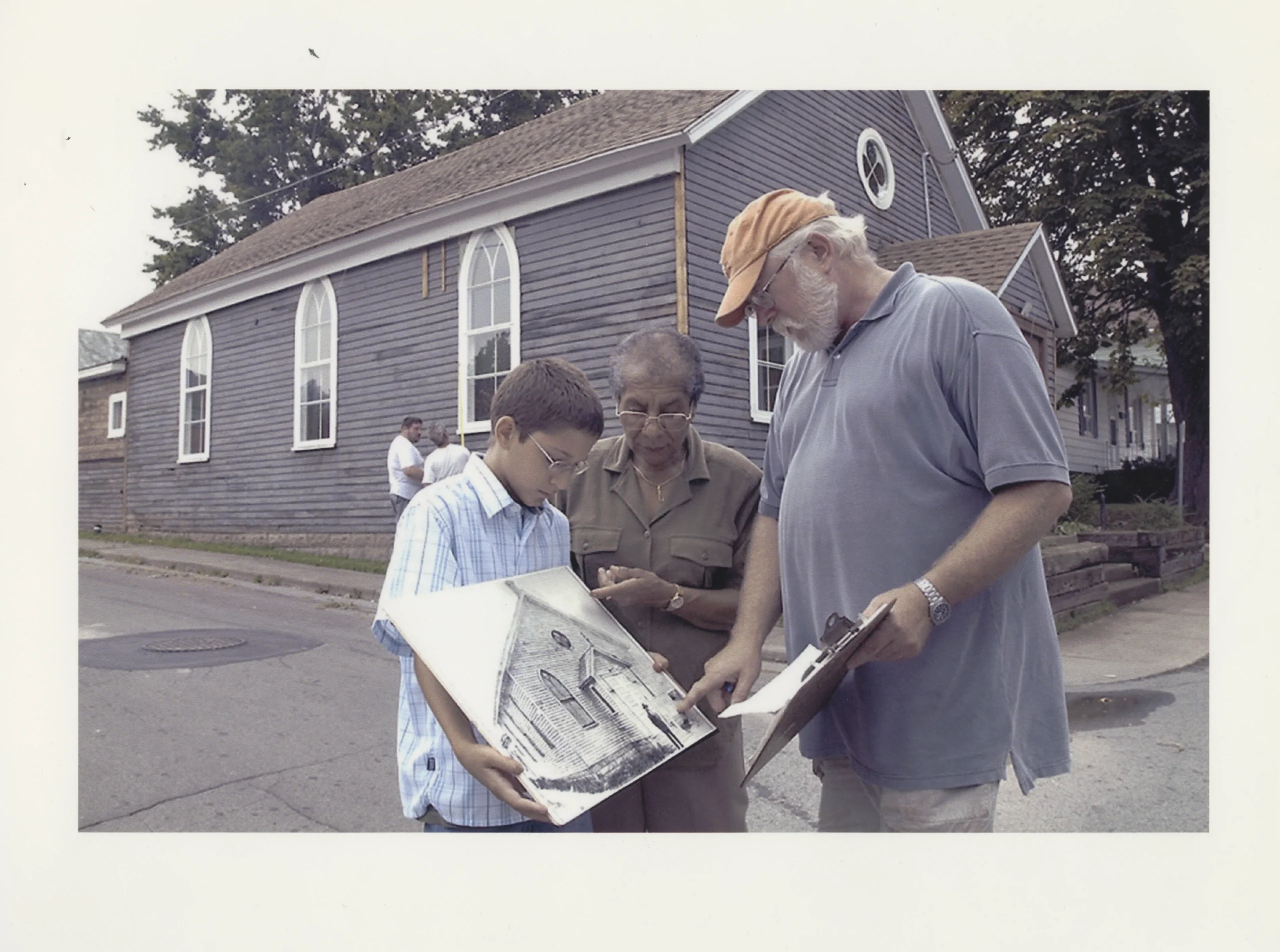 Photo couleur de trois personnes se tenant devant une petite église en bois, qui tiennent et regardent des plans architecturaux de l’église qui se trouvent dans une grande chemise.