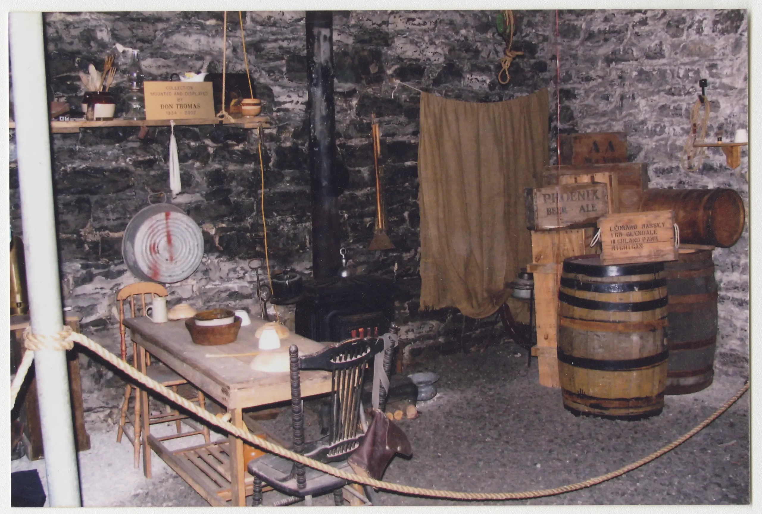 Photo couleur d’un coin d’une cave aux murs en briques, meublé avec une petite table et des chaises en bois, la table dressée avec des ustensiles de repas. La pièce contient aussi un poêle noir à long tuyau, un morceau de tissu suspendu à une corde, une grande marmite et quelques grands tonneaux et caisses en bois. Le mobilier est délimité par un cordon de sécurité.