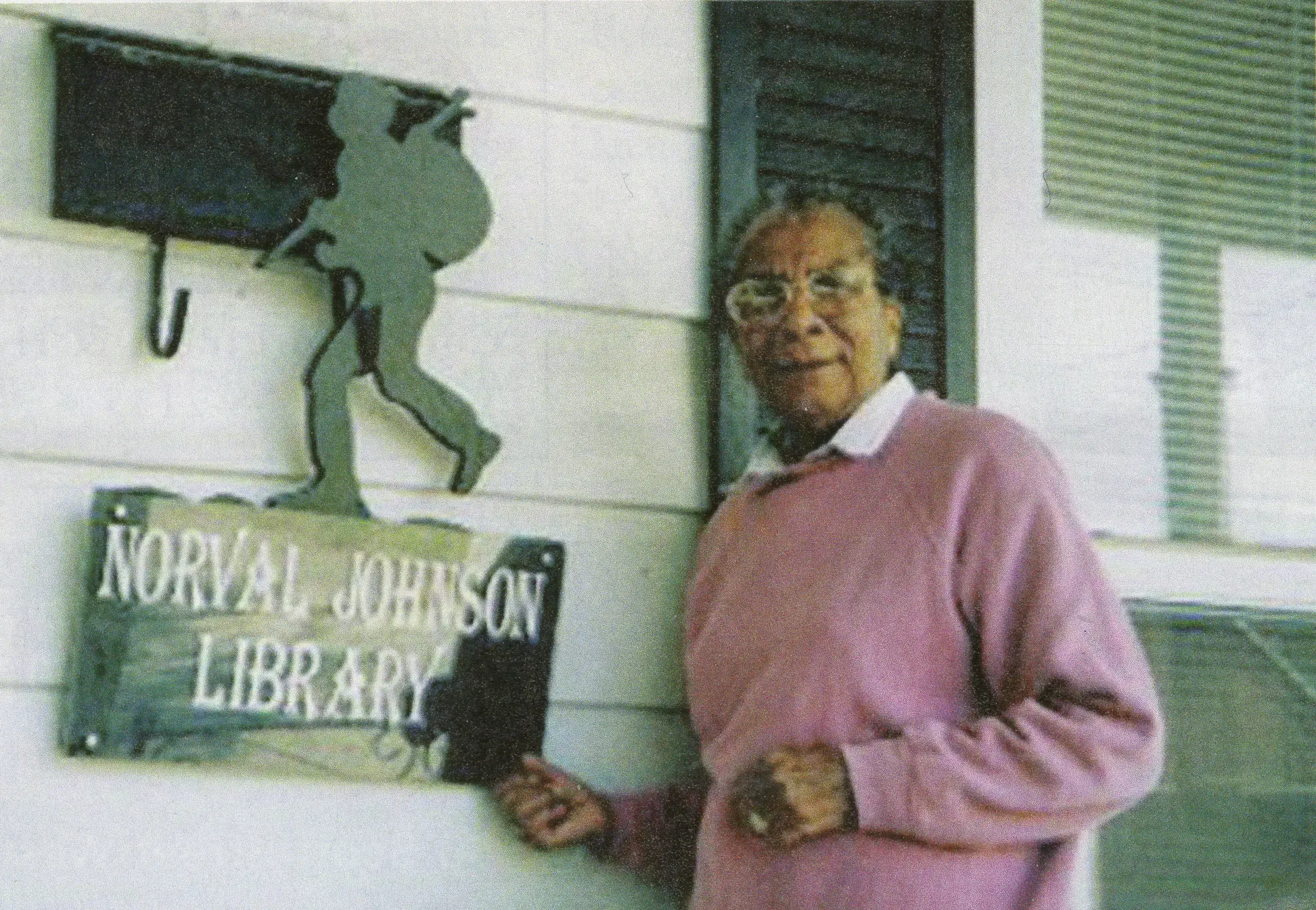 Photo couleur d’une femme âgée noire vêtue d’un chandail rose se tenant devant l’entrée d’un bâtiment à revêtement en bois blanc. Elle tient le coin d’un panneau fixé au mur, orné d’une silhouette d’homme qui marche en portant un baluchon, et sur lequel est inscrit « Bibliothèque Norval Johnson ».