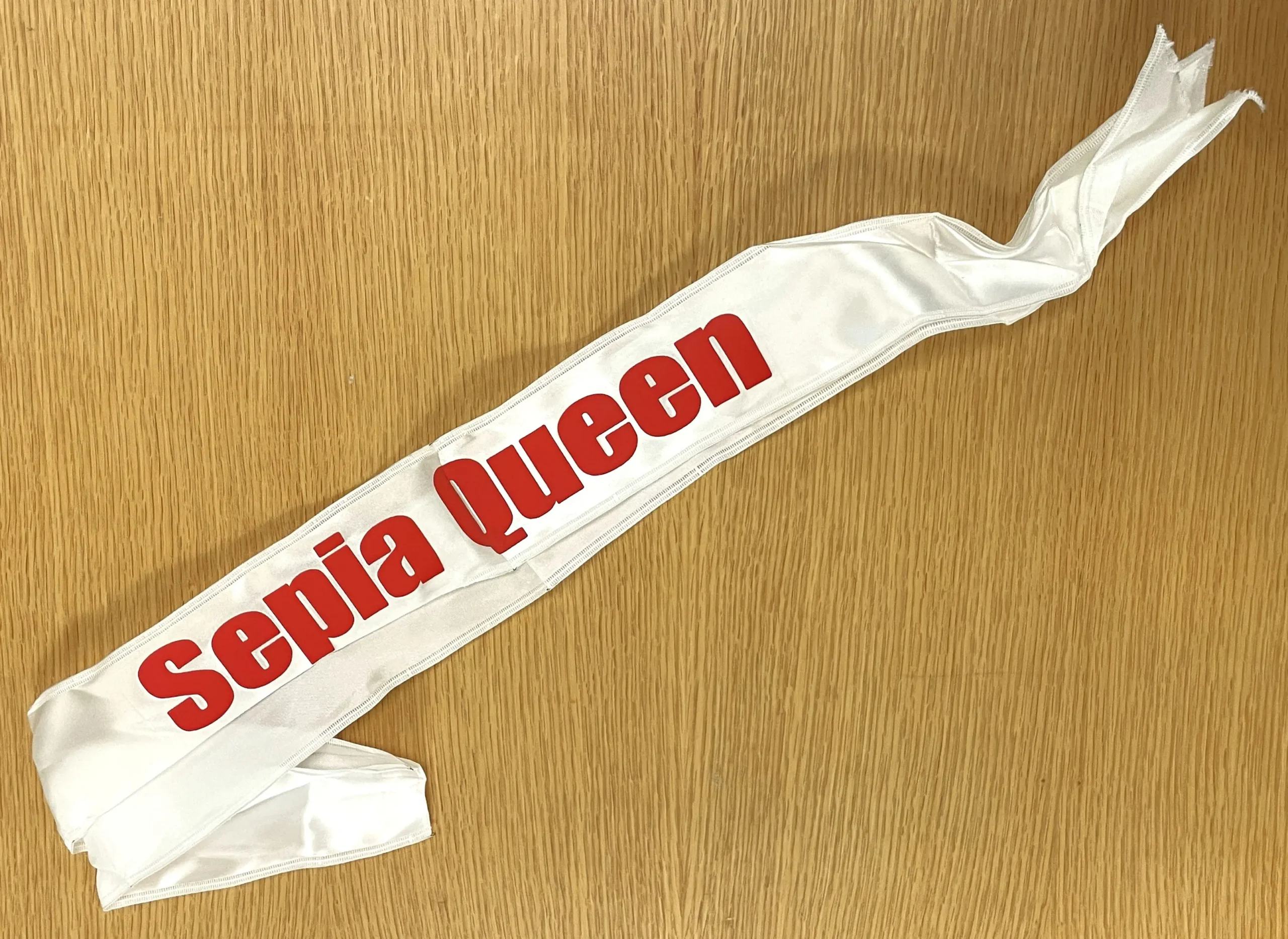 Photo couleur d’une banderole de soie blanche sur laquelle sont imprimées des lettres rouges formant les mots « Sepia Queen ».La collection comprend la banderole de l’équipe de basketball « Sepia Queens » de Mme Morrison