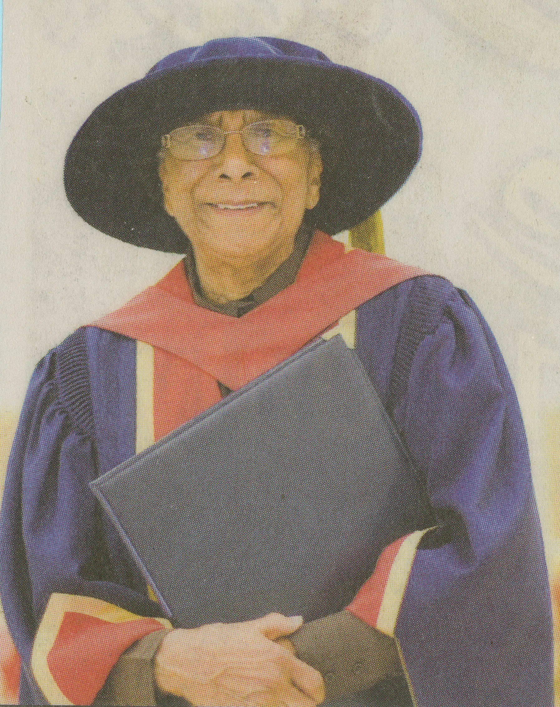 Photo couleur d’une femme âgée noire souriante portant une toge bleu, blanc et rouge, un mortier bleu, et tenant un porte-document en cuir noir.