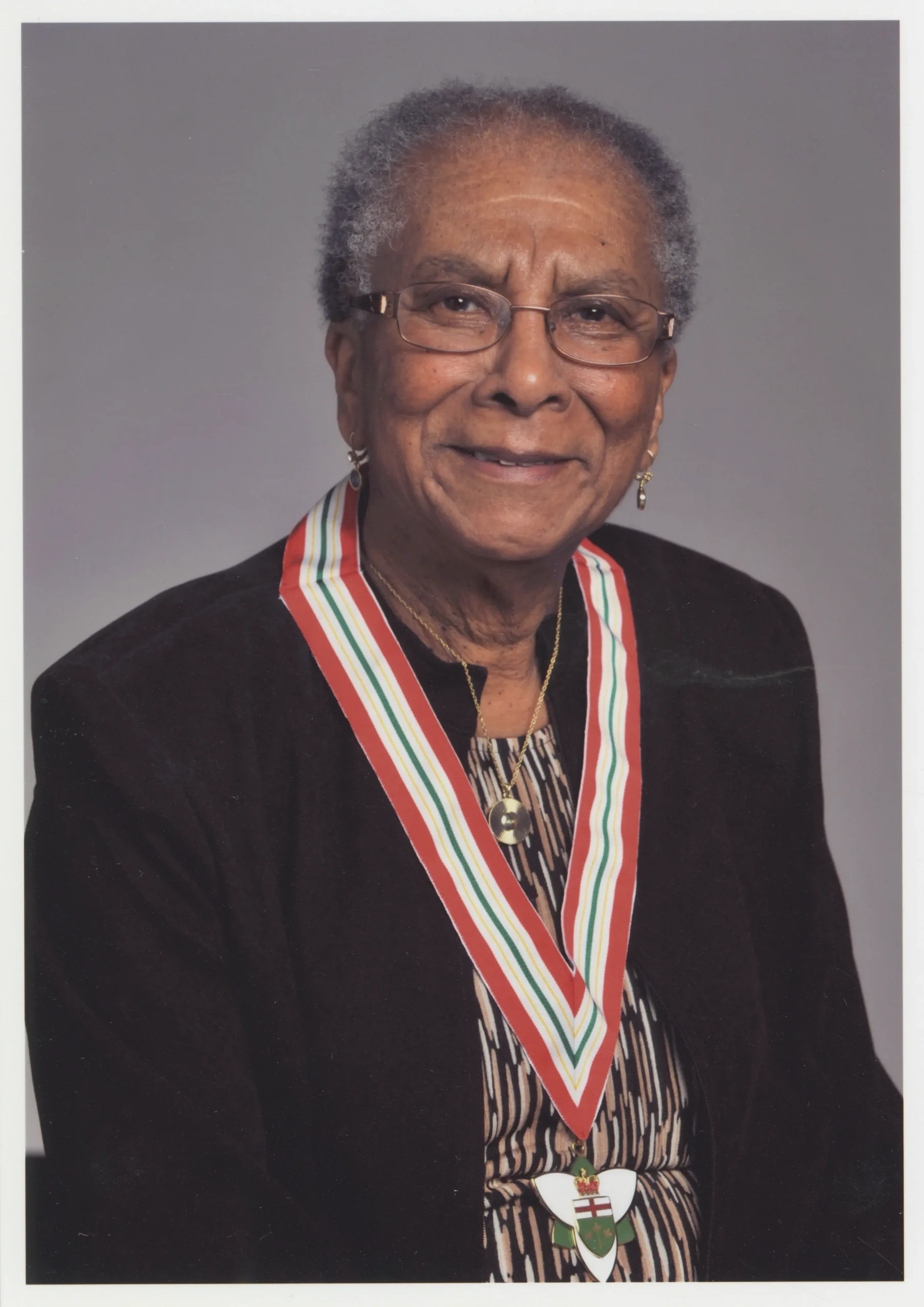 Portrait studio couleur format buste d’une femme noire âgée souriante, aux cheveux gris courts et portant des lunettes, arborant autour du cou une médaille portant l’insigne de la province de l’Ontario.
