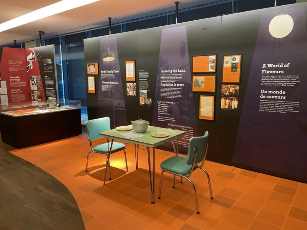 Colourful exhibit space featuring a green kitchen table and chairs. Wall panels are covered with text and images, against a black and purple background. The floor is covered with an orange tile pattern.