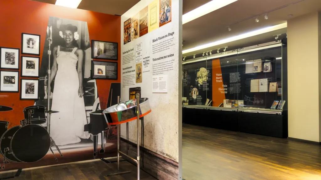 Colourful exhibit space featuring wall panels and glass display cases, displaying historical objects, books, images and texts. A steelpan drum can be seen in the foreground.