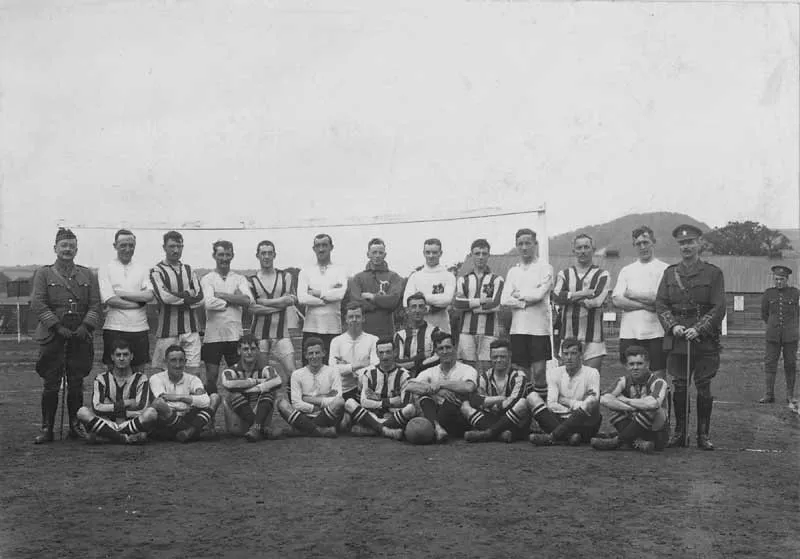 Black-and-white photo of a men’s soccer team posing in two rows. Their uniforms alternate from plain white jerseys to striped jerseys, and they’re flanked by two soldiers in uniform.