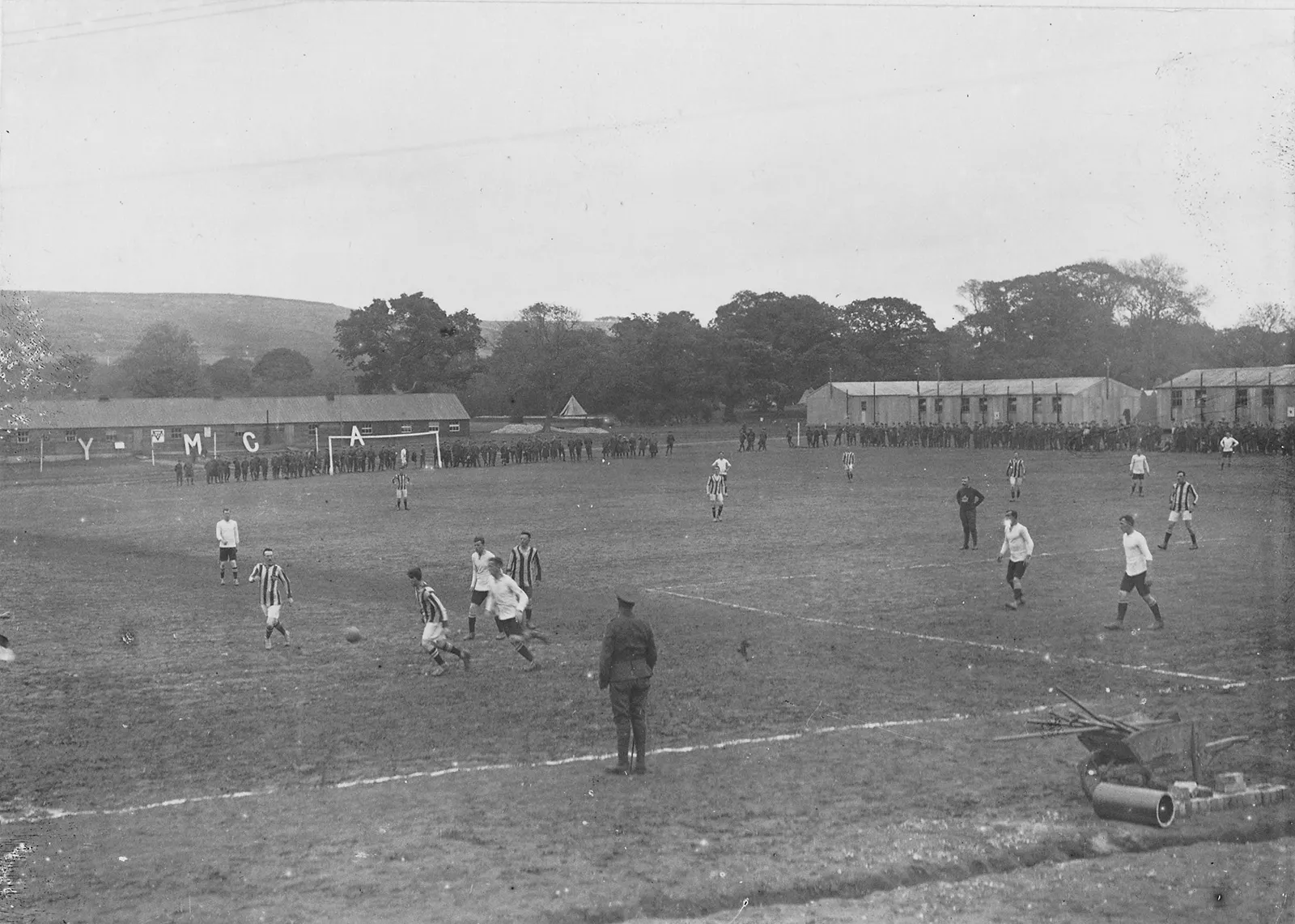 Black-and-white photo of soldiers watching a soccer game in a large open field. A player kicks the ball to the corner, attempting to make a play upfield. Soldiers in uniform are standing on the sidelines.