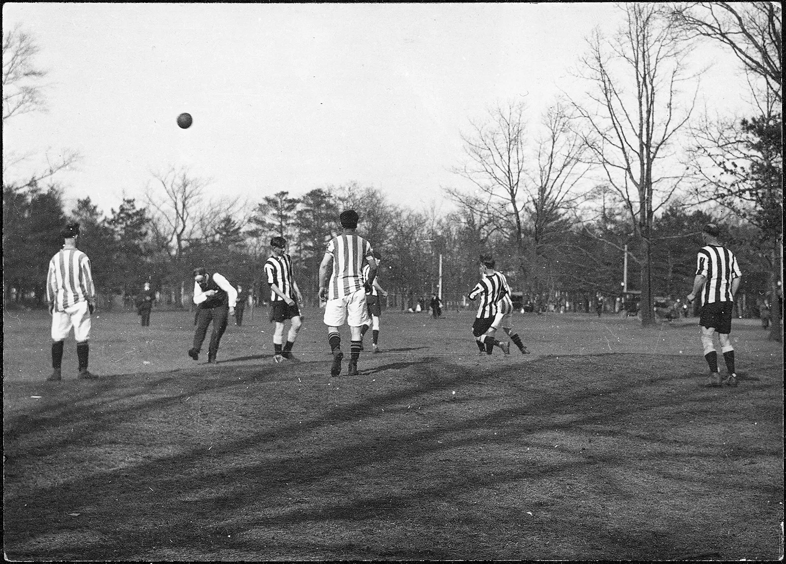 Black-and-white photo of men wearing striped shirts engaged in a game of soccer on a field. A defender appears to be kicking a ball upfield, and the referee, who is wearing a waistcoat, quickly moves out of the way to avoid being hit.