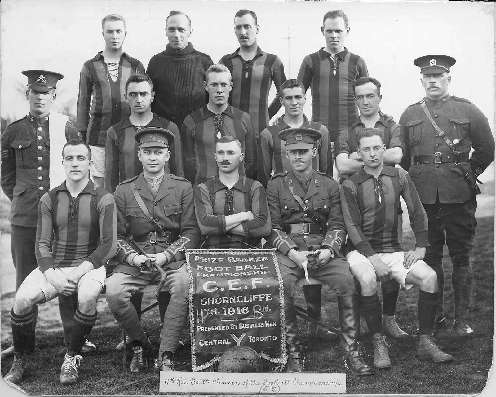 Black-and-white photo of a men’s soccer team with most players in striped jerseys but some in military fatigues. Members are arranged in three rows with a prize banner and a soccer ball front and centre.