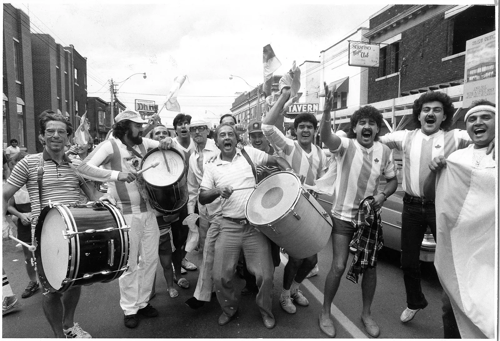 Black-and-white photo of Argentine fans celebrating on the street by banging drums and singing as Argentina wins the FIFA™ World Cup. Fans are waving the Bandiera Nacional, which is another name for the Argentinian flag.