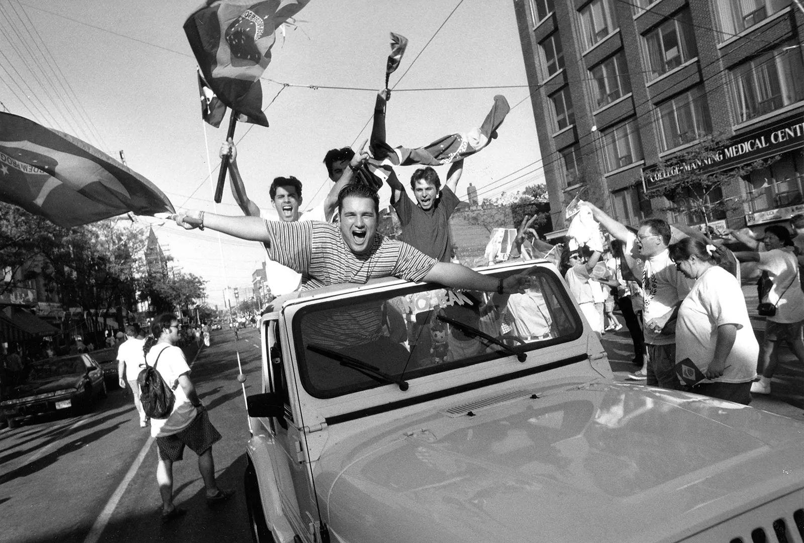 Black-and-white image of an open-top car driving toward the camera, carrying celebrating fans who cheer and wave their arms and flags. Crowds are also assembled on the street.