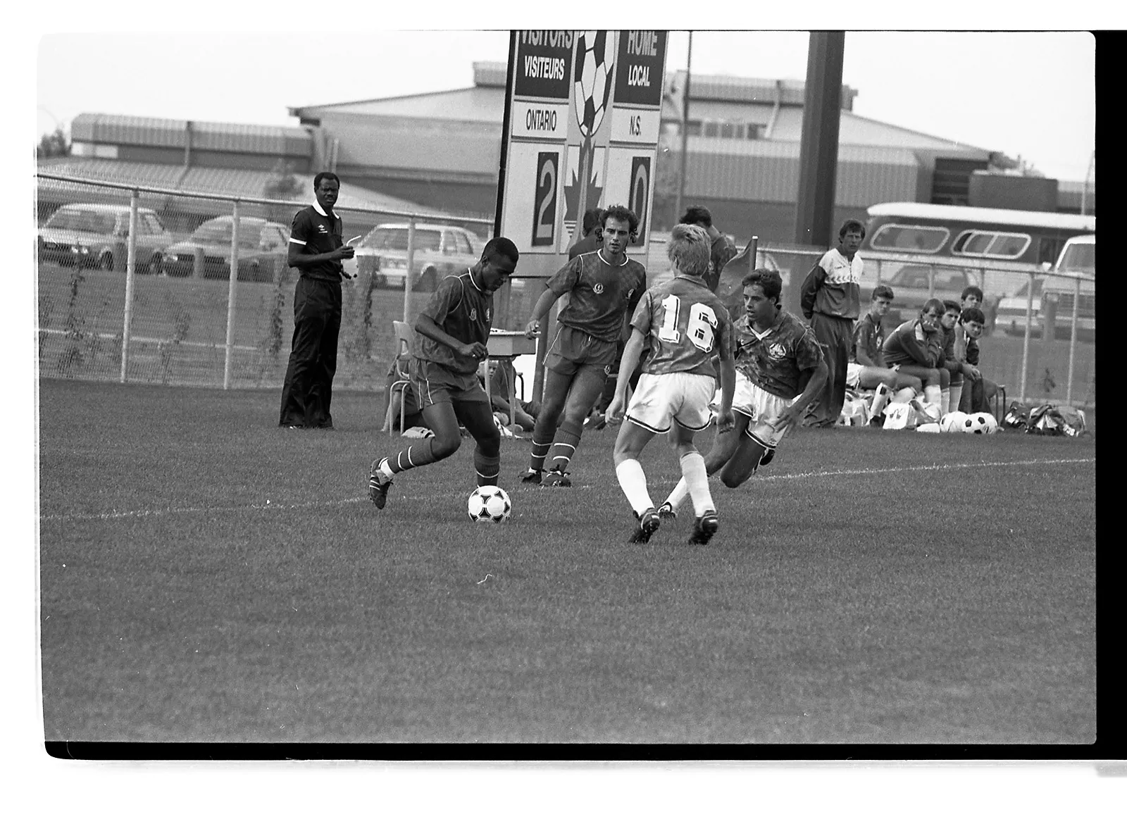 Black-and-white photo of a soccer player dribbling the ball near the by-line, attempting to get past a defender on the opposing team.