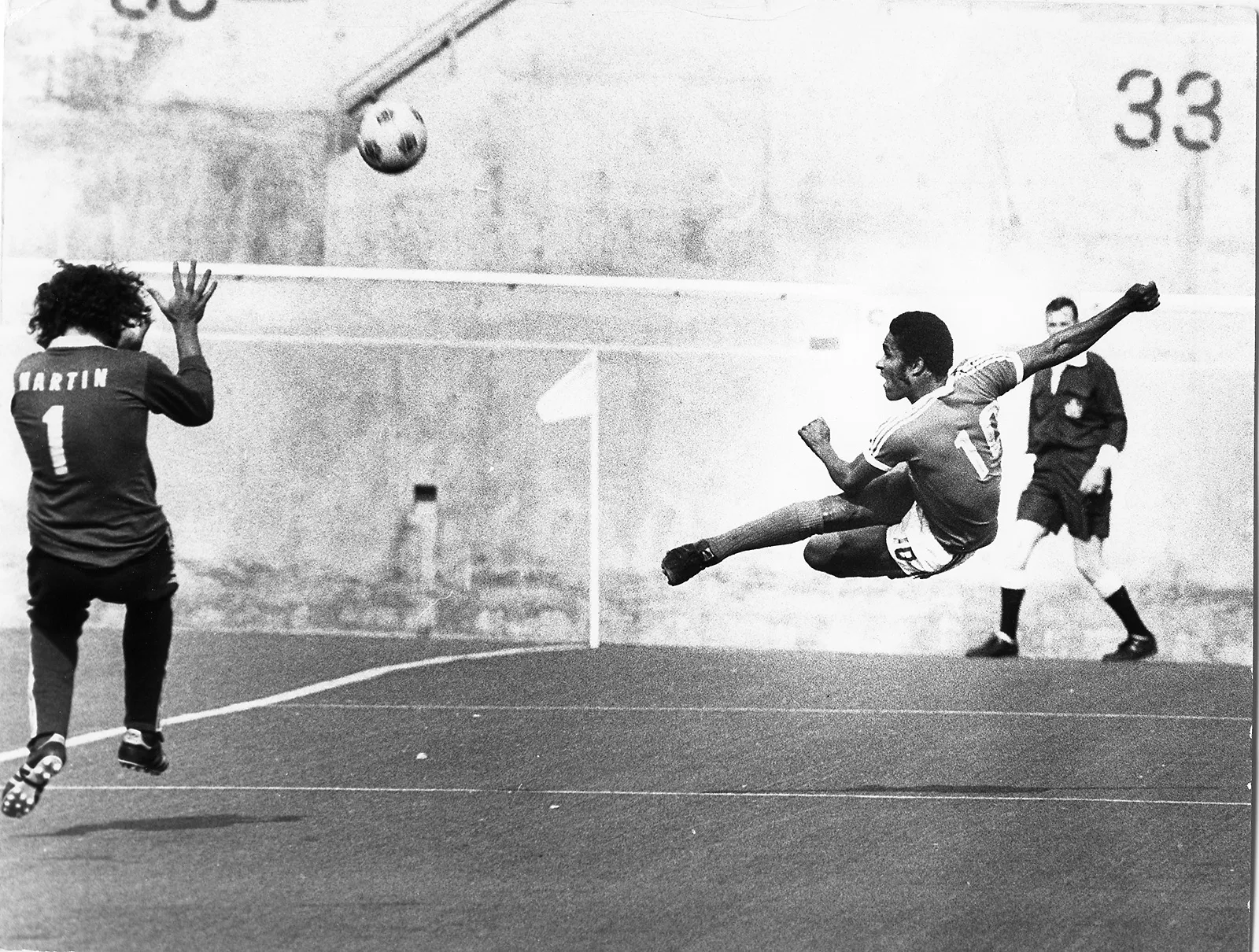 Black-and-white photo of a Black soccer player delivering an acrobatic kick, directing the ball towards the goal. The goalie dives to make an attempted save.