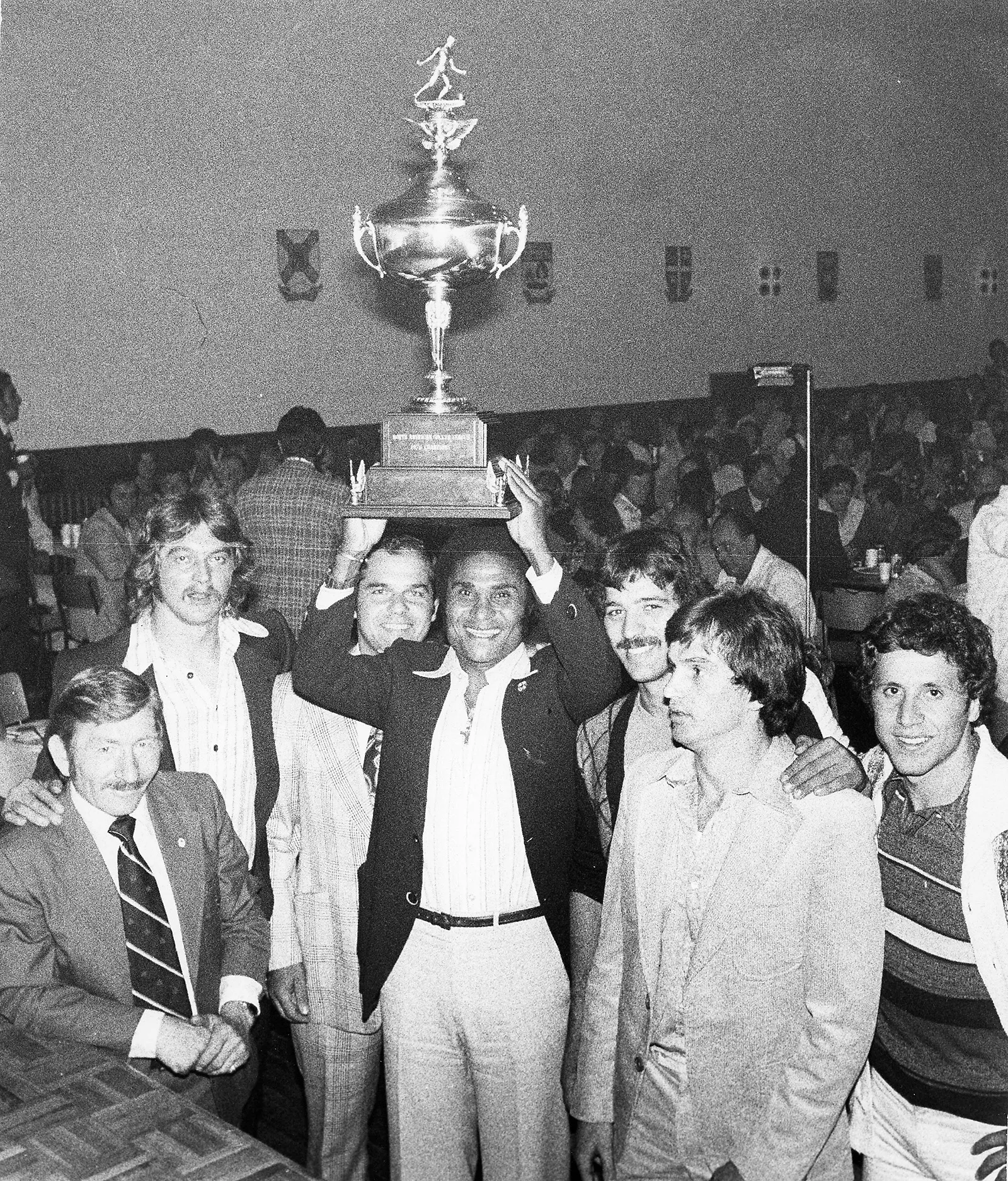 Black-and-white photo of a Black man lifting a large trophy over his head in a room full of people. His teammates and coaches stand by his side and smile at the camera.