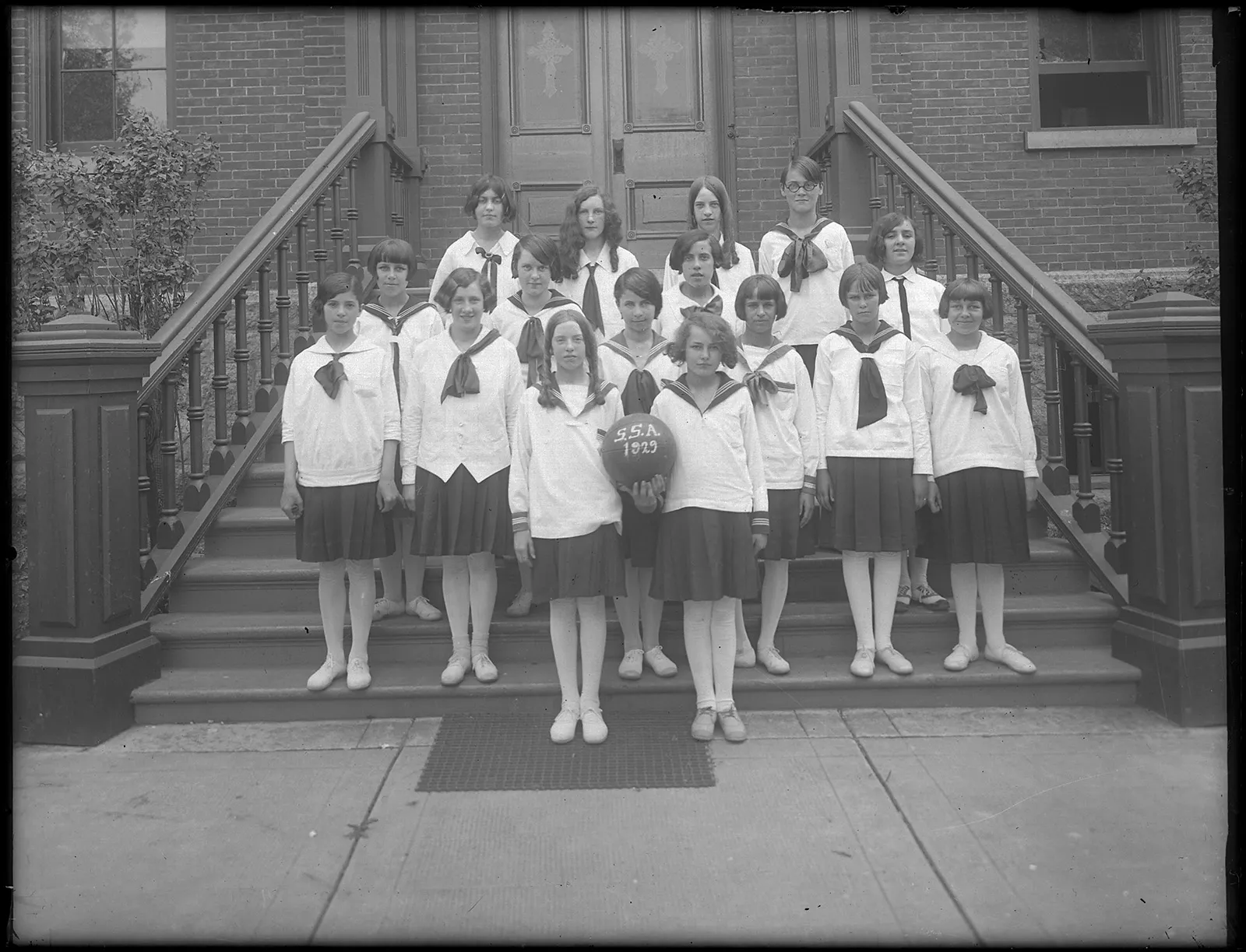 Black-and-white portrait of an all-girls soccer team wearing light-coloured, long-sleeved tops with scarves and dark skirts. The girls are standing on the steps of what appears to be a school. The two girls standing at the front of the group hold a ball bearing the initials SSA, which might represent an academic team.