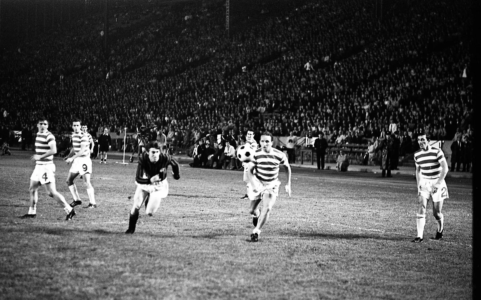Black-and-white photo of a soccer player from Celtic and a player from Milan chasing down a ball on an outdoor stadium soccer field.