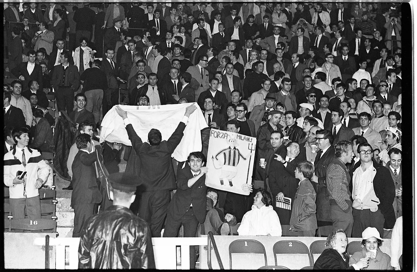 Black-and-white photo of fans packed in the bleachers at a soccer game. A fan holds a poster with an image of a devil wearing a Milan jersey, a popular mascot of the team.