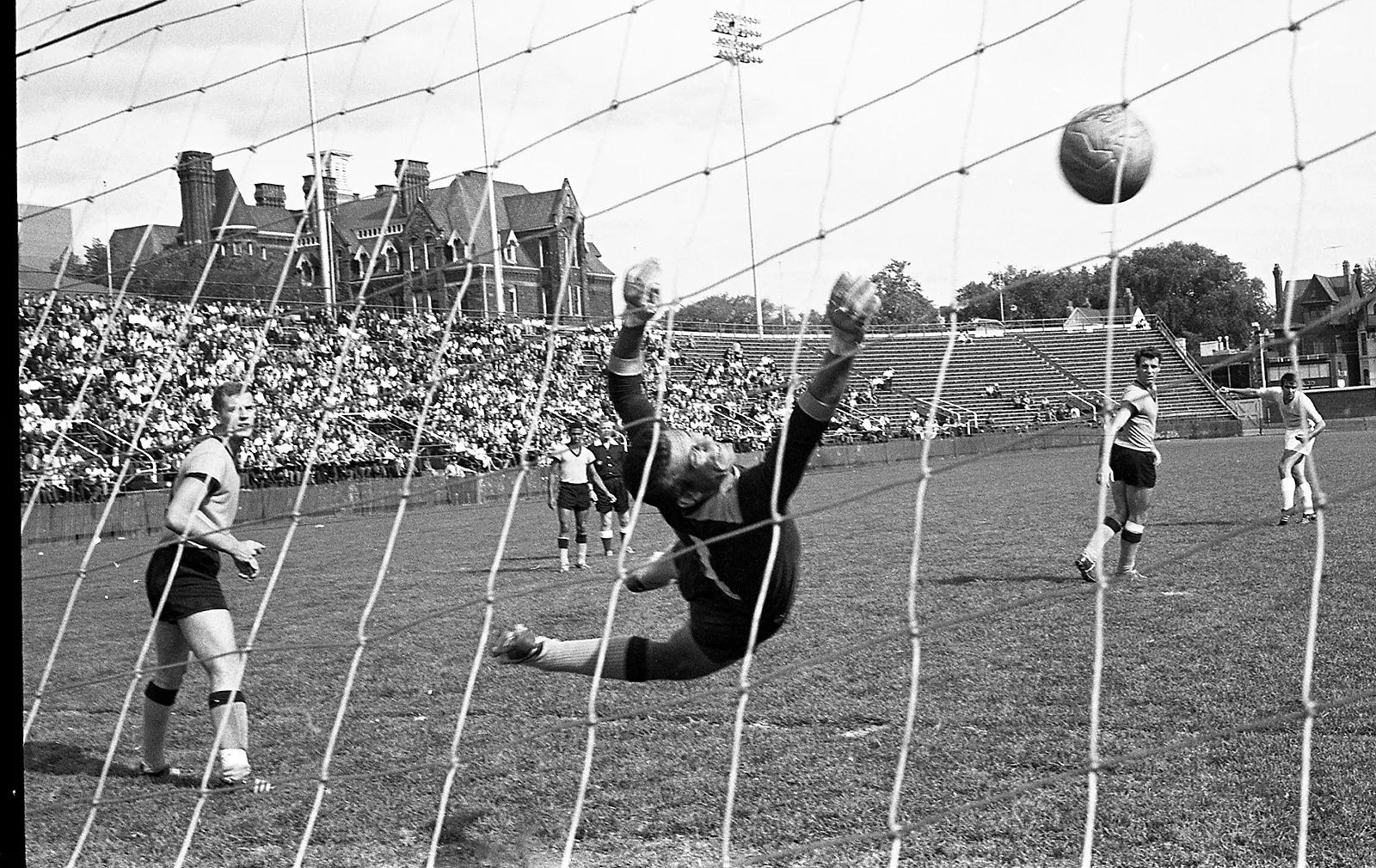 Black-and-white photo of a goalie fully stretched out, attempting to make a save, seen from behind the net of the goal. A large crowd of people in the stands, along with other players on the field, watch the goalie to see if he makes the save.