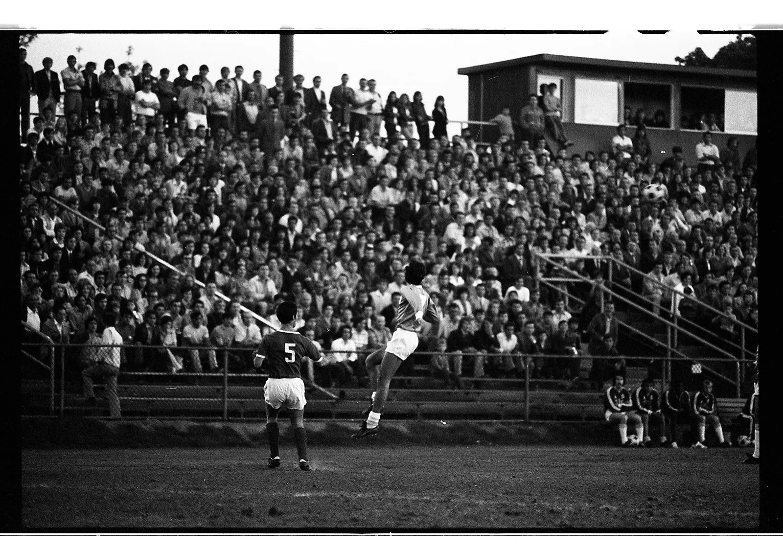 Black-and-white photo of a player jumping in the air as a ball goes through the air. A large crowd of spectators watches from the bleachers, a row of them standing at the top of the stands as there are no seats left for them to sit.