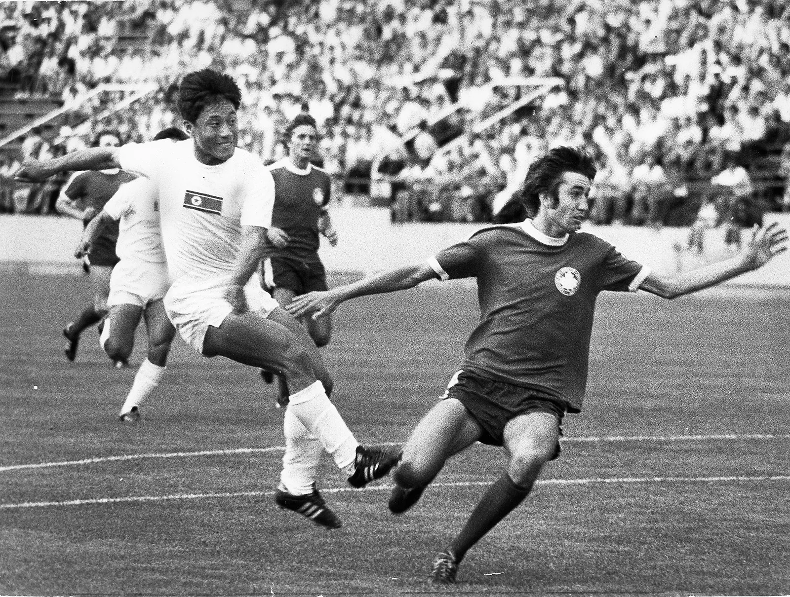 Black-and-white photo of a North Korean player kicking a ball towards the net as a Canadian player attempts to make a block. The stands are packed with spectators in the background.