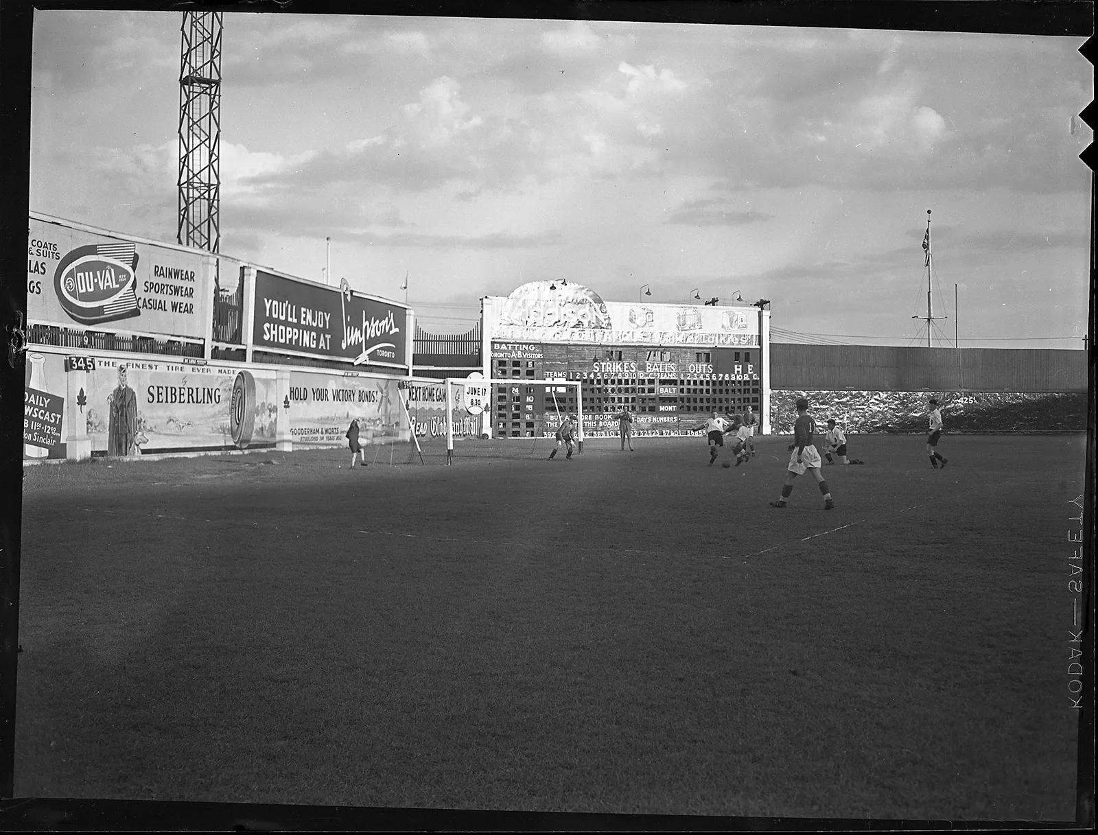 Black-and-white photo of a soccer game on an outdoor stadium field. Two players in the box fight for position to take control of the ball. Behind the goal is the outfield wall of the baseball field, with billboards displaying advertisements, including one promoting victory bonds.