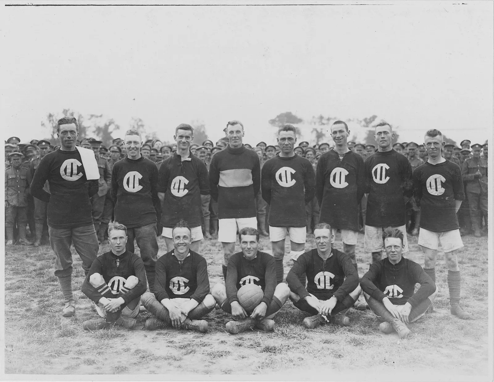 Black-and-white photo of a men’s soccer team, with members arranged in two rows. A large group of military servicemen can be seen standing behind them, in the distance.