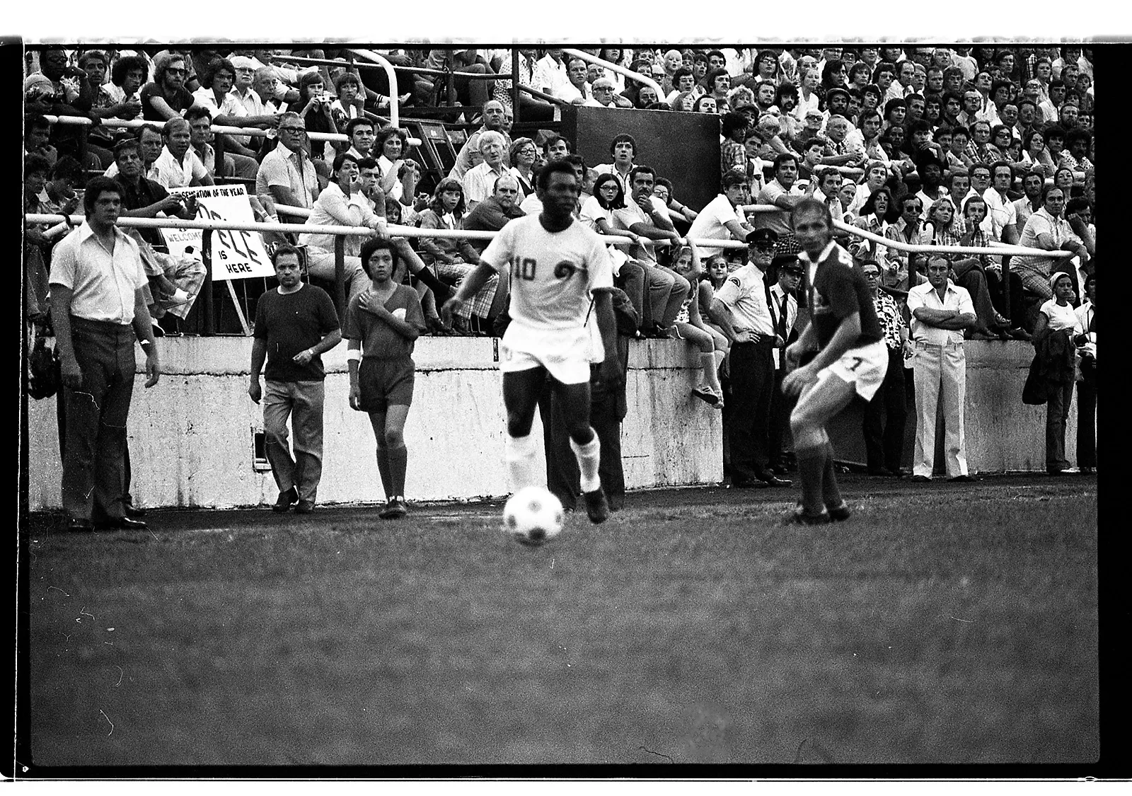 Black-and-white photo of a Black soccer player running out of a tunnel beneath the stands in a stadium, waving his hand in the air. The crowd of people in the stands behind cheer him on.