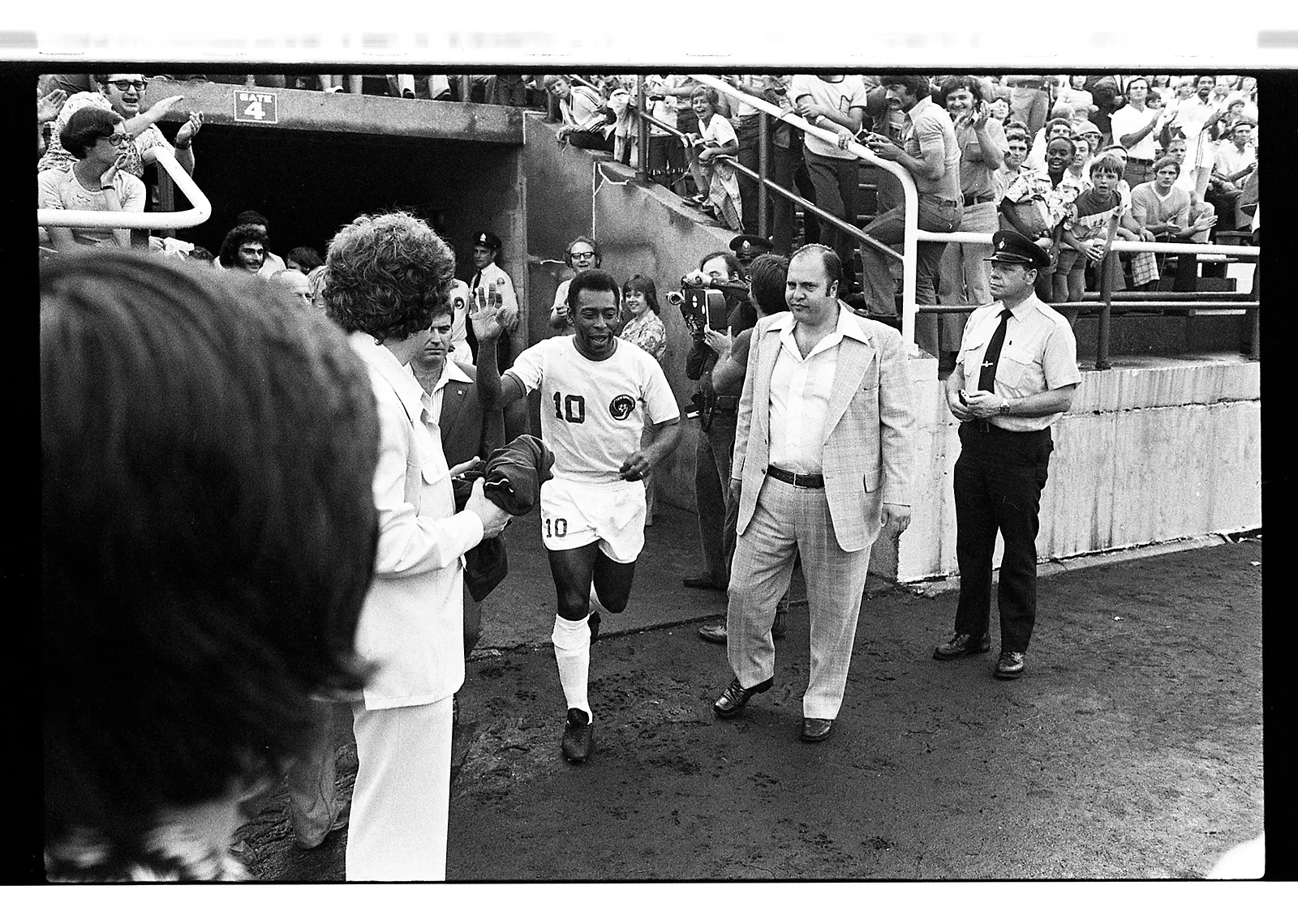 Black-and-white photo of a Black soccer player running out of a tunnel beneath the stands in a stadium, waving his hand in the air. The crowd of people in the stands behind cheer him on.