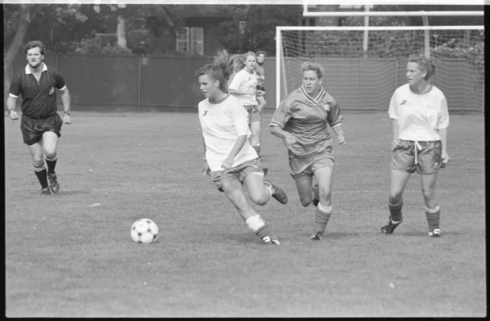 Black-and-white photo of a soccer player kicking a ball towards the goal as a player on the other team gives it her all and chases the opposing player down.