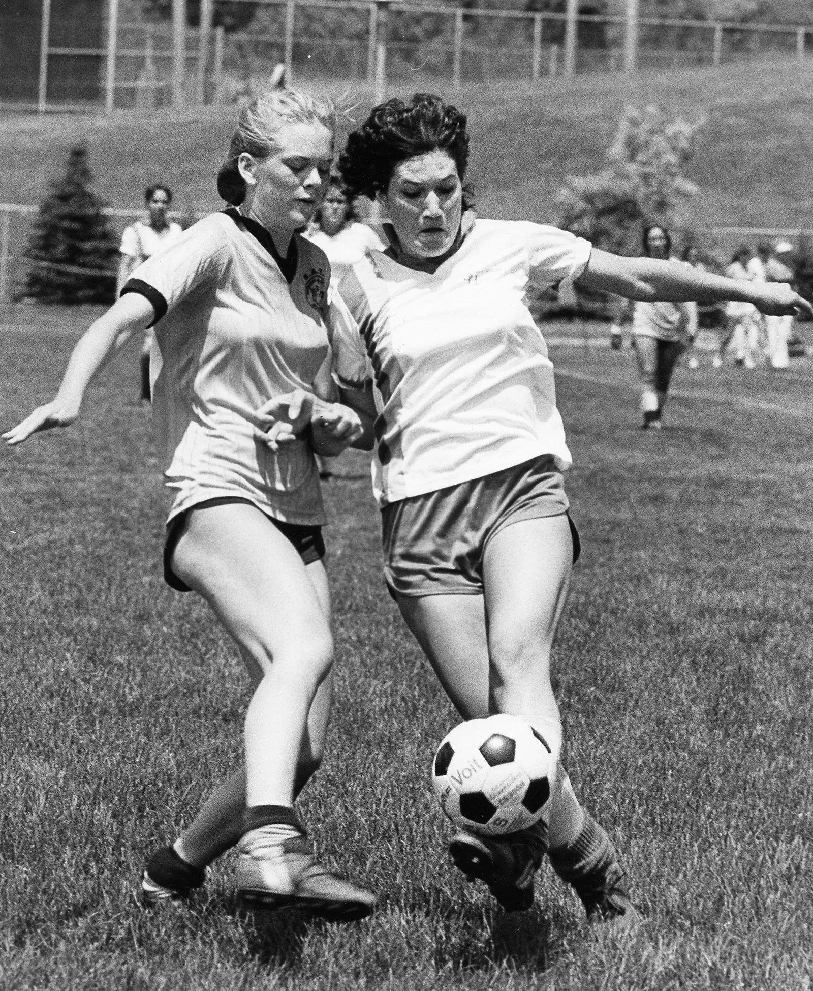 Black-and-white photo of two women soccer players. One seems to be flicking the ball around her opponent, as the defender uses her strength to keep her to the outside.