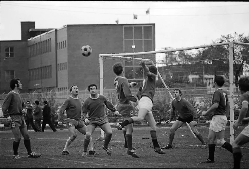 Black-and-white photo of a ball flying across the goal from a corner kick. There is a mix of bodies in the 6-yard box as players try to get a touch on the ball. A multi-story institutional-looking building can be seen in the background.
