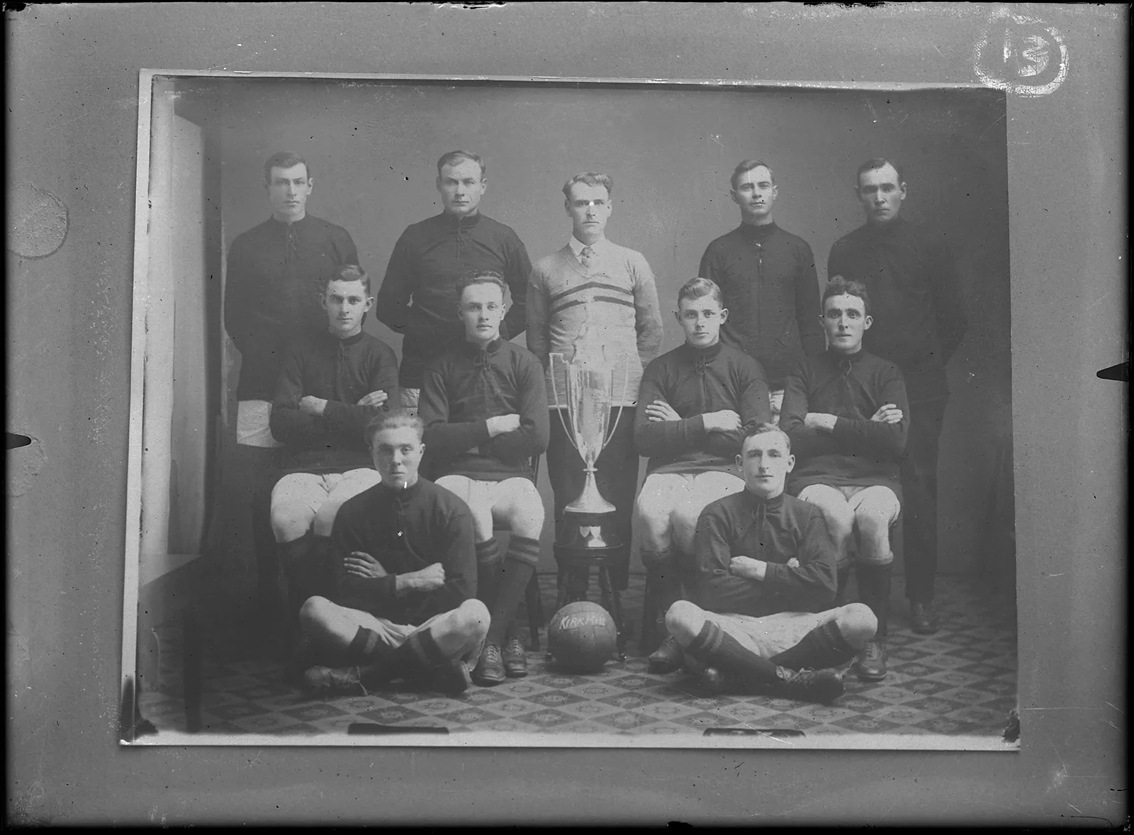 Black-and-white photo of a team portrait of an amateur men’s soccer team. The players are arranged in three rows and proudly surround a trophy they have just won.