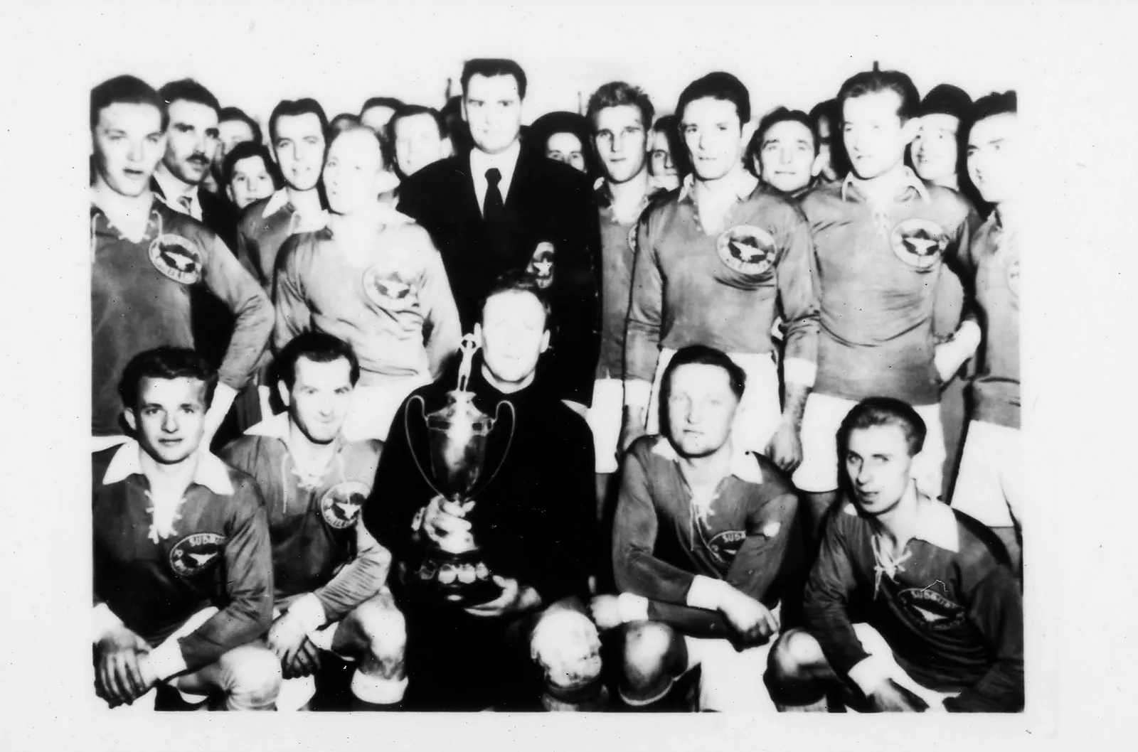 Black-and-white photo of a soccer team. The players have an insignia on their jerseys. The team smiles at the camera as they proudly display their trophy. There appear to be fans crowded in the background.