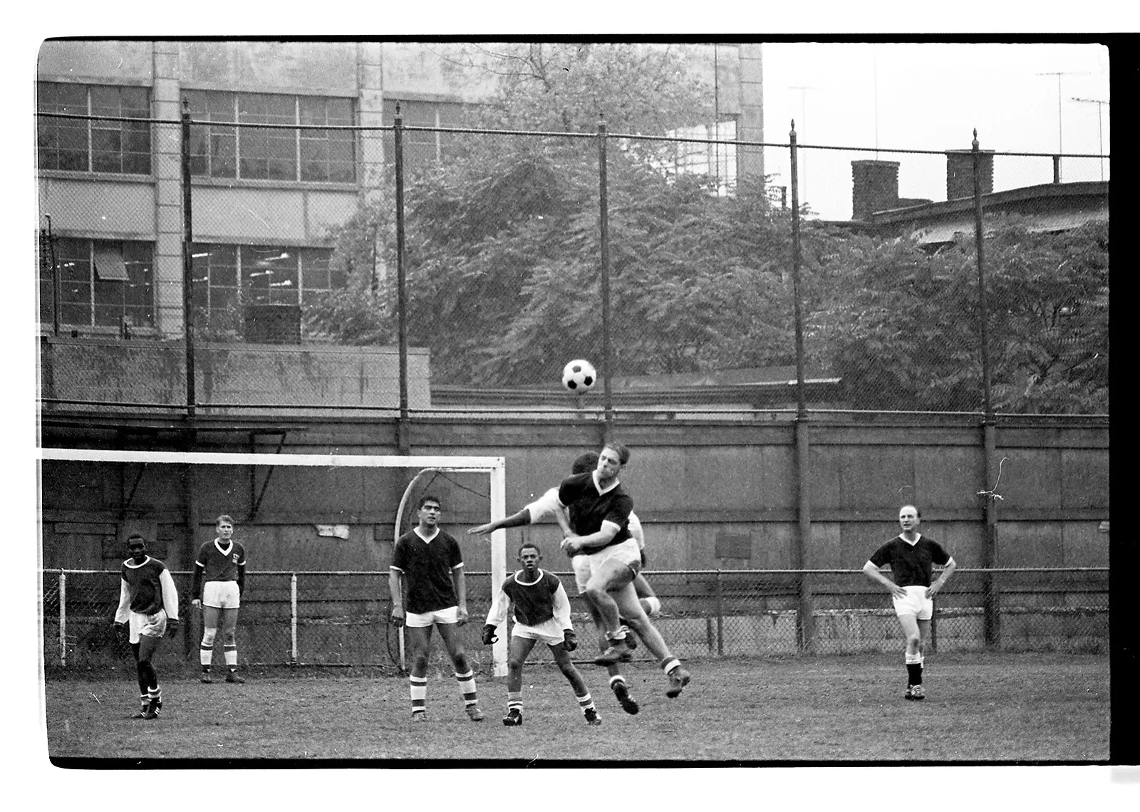 Black-and-white photo of two soccer teams engaged in a game on an outdoor field. Two players from opposing teams battle it out in the air for an incoming ball. The other players wait to see where the next play will happen.